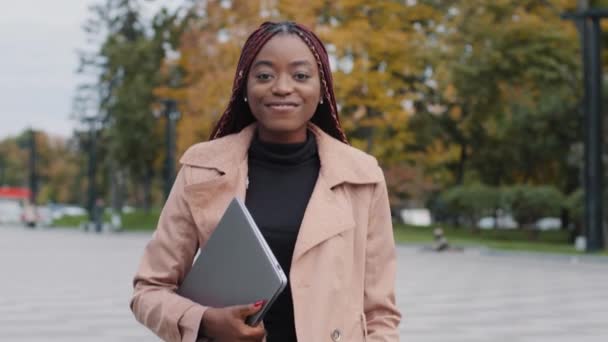 Beautiful smiling millennial African American female manager with laptop, successful businesswoman or student walking downtown in casual stylish autumn clothing. Happy young lady looking at camera  - Imágenes, Vídeo