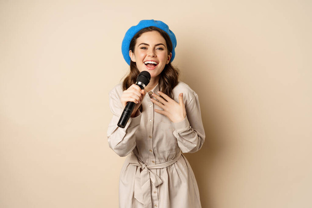 Portret van vrolijke stijlvolle vrouw optredend, zingend met microfoon, poserend tegen beige achtergrond - Foto, afbeelding