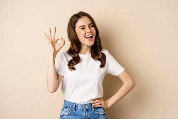 Stylish young woman smiling satisfied, showing okay, ok sign in approval, recommending, standing over beige background - Foto, imagen