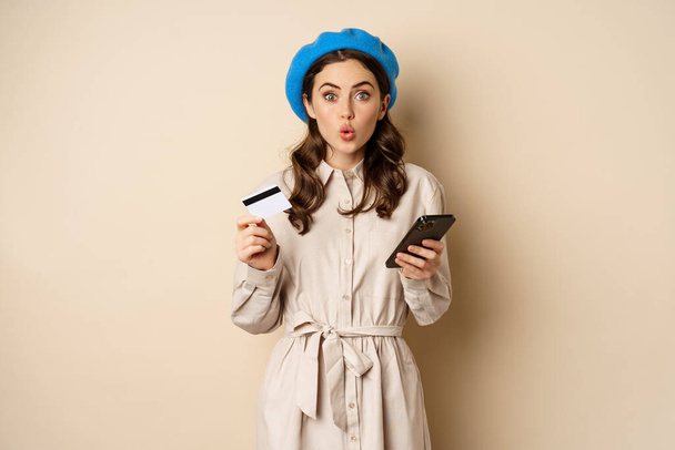 Portrait of girl looking excited, say wow, holding credit card and mobile phone, standing over beige background - Fotografie, Obrázek