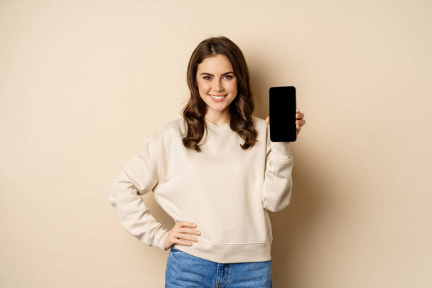 Stylish smiling woman showing smartphone screen, mobile app interface, standing over beige background - Zdjęcie, obraz