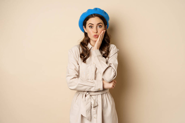 Image of stylsh beautiful woman looking surprised, shocked reaction at camera, posing in trench coat against beige background - Fotografie, Obrázek