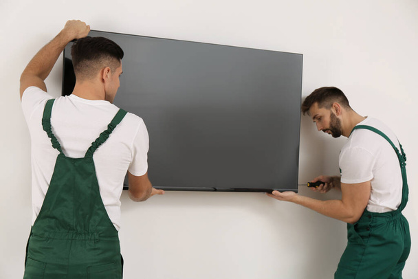 Professional technicians with screwdriver installing modern flat screen TV on wall indoors - Foto, imagen