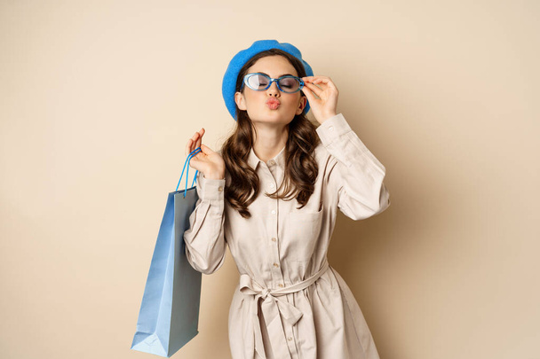 Стильна щаслива дівчина на покупках. Портрет сучасної жінки з сумкою в магазині, сміється і посміхається задоволений, купуючи собі подарунок, стоячи над бежевим тлом
 - Фото, зображення
