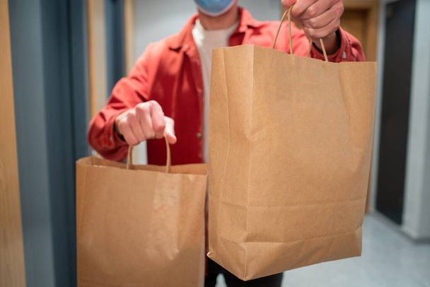 Delivery man in protective mask holding paper bag with food in the entrance. The courier gives the box with fresh vegetables and fruits to the customer - Fotografie, Obrázek