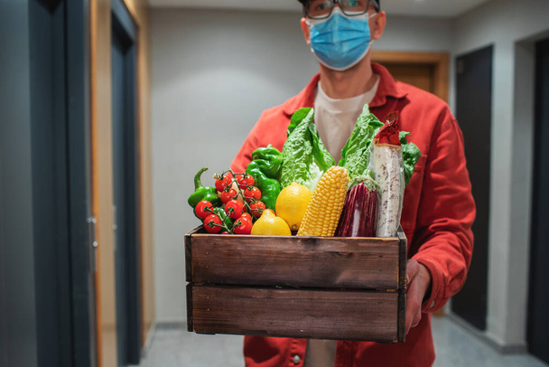 Доставщик в защитной маске держит бумажный пакет с едой на входе. Курьер дает коробку со свежими овощами и фруктами клиенту - Фото, изображение
