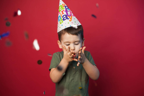 Caucasian boy in paper hat blowing confetti isolated over red background. Holiday celebration. Holiday, birthday. Holiday decoration. Present birthday. Red - Fotoğraf, Görsel