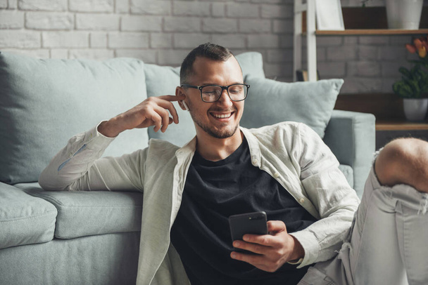 Portrait d'un homme souriant utilisant un téléphone portable et des écouteurs sans fil assis sur le sol à la maison. - Photo, image
