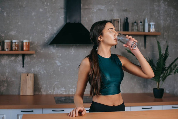 Blanke vrouw die in de huiskeuken staat en vers water drinkt na het sporten. Dieet, gezonde levensstijl, gewichtsverlies concept. Schoonheidsconcept. Huis - Foto, afbeelding