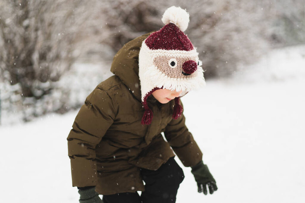 Cute little boy in funny winter hat walks during a snowfall. Outdoors winter activities for kids. - Фото, изображение