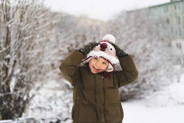 Cute little boy in funny winter hat walks during a snowfall. Outdoors winter activities for kids. - Foto, imagen
