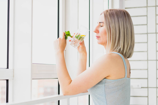 Молода щаслива жінка стоїть біля віконної склянки освіжаючого напою з лимоном і м'ятою. Здоровий спосіб життя, омолодження
 - Фото, зображення