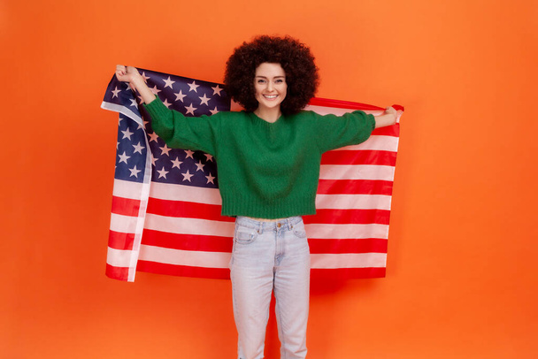 Happy positive woman with Afro hairstyle wearing green casual style sweater standing with raised arms, holding USA flag, celebrating national holiday. Indoor studio shot isolated on orange background. - Photo, Image
