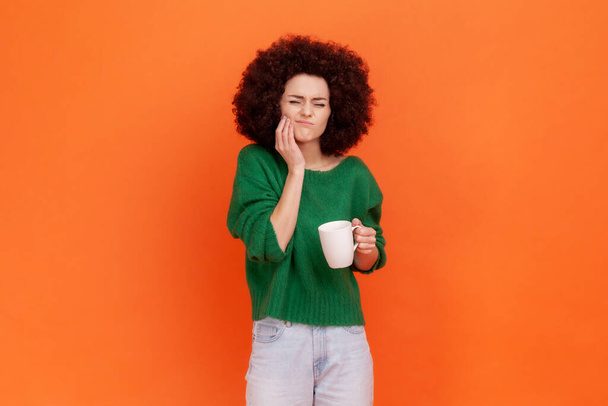 Sad sick woman with Afro hairstyle wearing green casual style sweater has teeth pain after drinking hot or cold beverage, dental injury. Indoor studio shot isolated on orange background. - Photo, image