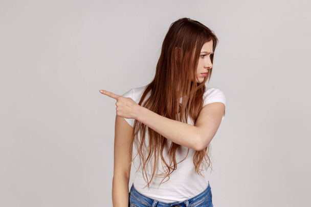 Portrait of resentful upset attractive woman turning away with disgust and ordering to leave, showing way out, feeling betrayed, wearing white T-shirt. Indoor studio shot isolated on gray background. - Photo, Image