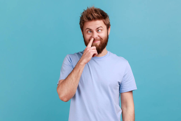 Misconduct, bad manners. Portrait of crazy bearded man picking nose and sticking out tongue with dumb comical expression, fooling around. Indoor studio shot isolated on blue background. - Photo, Image