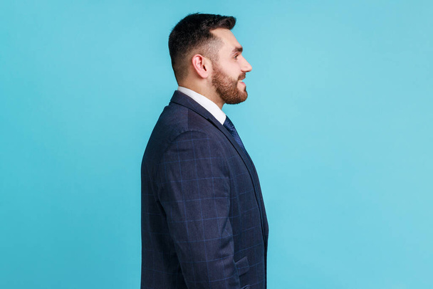 Profile portrait of young adult handsome bearded businessman wearing official style suit looking ahead with toothy smile, optimism, success. Indoor studio shot isolated on blue background. - Zdjęcie, obraz