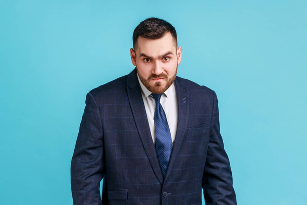 Angry handsome businessman wearing official style suit looking at camera, having aggressive facial expression, expressing negative, hatred. Indoor studio shot isolated on blue background. - Fotoğraf, Görsel