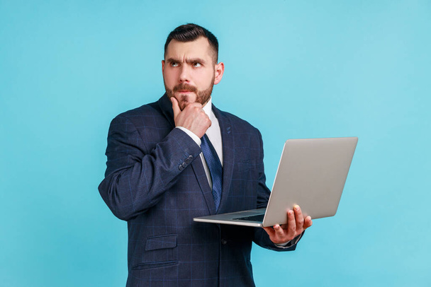 Portrait of pensive smart businessman wearing dark official style suit holding laptop and thinking about new project, holding his chin. Indoor studio shot isolated on blue background. - Foto, Bild