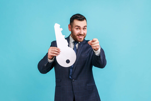 Happy man with beard wearing dark official style suit big paper key and pointing to camera with toothy smile, house purchase concept. Indoor studio shot isolated on blue background. - Photo, image