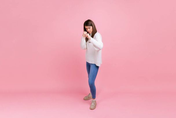 Full length of attractive angry female, expressing anger and aggression, boxing to camera, fighting, wearing white casual style sweater. Indoor studio shot isolated on pink background. - Foto, Imagem