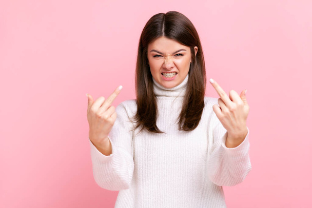 Portrait of angry aggressive female standing and shows fuck sign with both hands into the camera, wearing white casual style sweater. Indoor studio shot isolated on pink background. - Foto, immagini