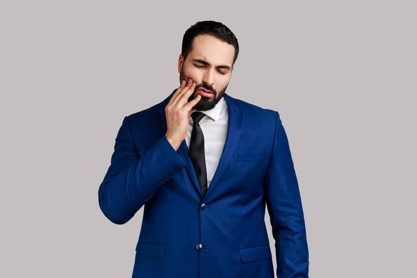 Unhealthy bearded man wincing in pain and touching sore cheek, suffering unbearable toothache, gum disease, wearing official style suit. Indoor studio shot isolated on gray background. - Фото, изображение