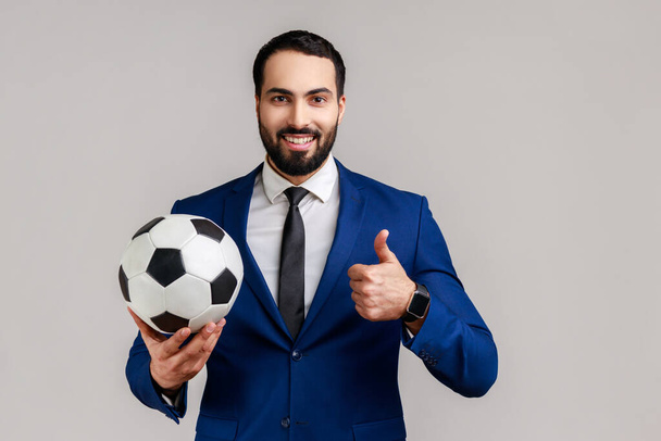 Satisfied bearded businessman holding soccer ball on his hand with smiling positive expression, showing thumb up, wearing official style suit. Indoor studio shot isolated on gray background. - Fotó, kép