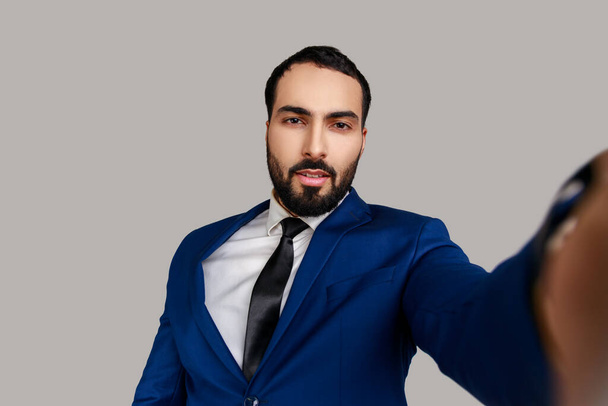 Portrait of handsome bearded man taking selfie, looking at camera with relaxed optimistic calm emotions, POV, wearing official style suit. Indoor studio shot isolated on gray background. - Foto, Imagem