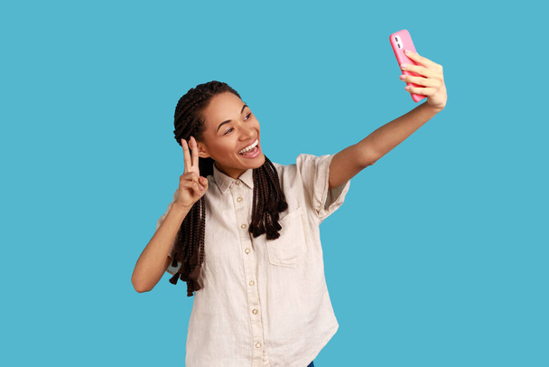 Glad woman with toothy smile, makes peace sign or V gesture at camera of cell phone, poses for making selfie or video call, wearing white shirt. Indoor studio shot isolated on blue background. - Foto, Imagem