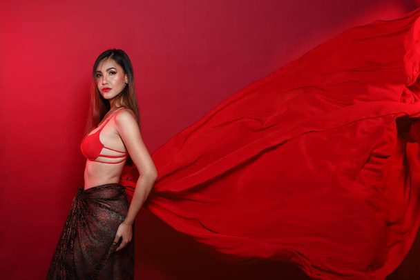 Portrait der jungen schlanken Transgender Asiatin trägt rotes Bikini-BH-Kleid, Schönes Mädchen flatternder Stoff wirft im Differenzstil eleganten Wellenwind in die Luft, roter Hintergrund - Foto, Bild