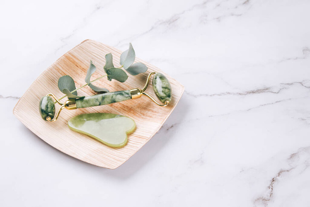 Jade facial roller and gua sha tool on white marble table background with copy space. Facial massage kit for lifting massage therapy made from natural stones, personal skin care anti-aging tools. Selective focus - 写真・画像