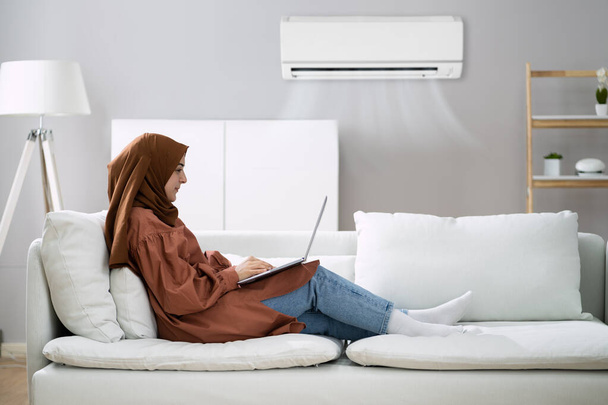 Woman Lying On Sofa Using Laptop Enjoying The Cooling Of Air Conditioner At Home - 写真・画像