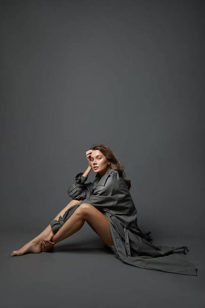 Hermosa mujer en traje de cuerpo beige y largo impermeable gris posa sentado en el suelo sobre un fondo gris. Largas piernas hermosas de un modelo de moda - Foto, imagen