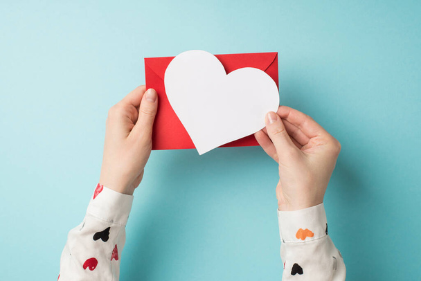 First person top view photo of saint valentine's day decorations female hands in white shirt holding red envelope and white paper heart on isolated pastel blue background with empty space - Photo, Image