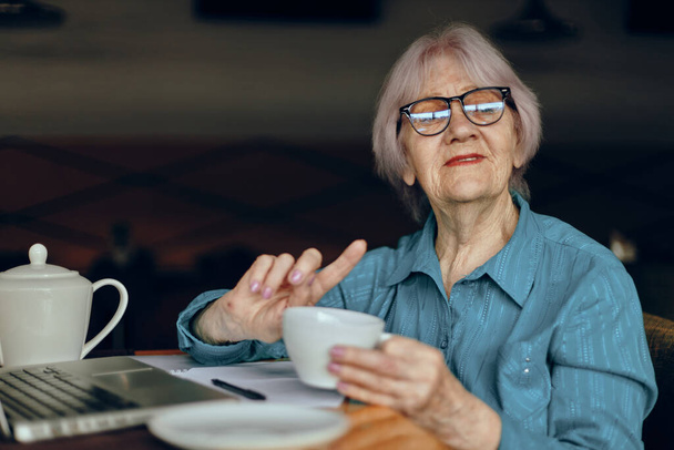 Portrait of an elderly woman with glasses sits at a table in front of a laptop Freelancer works unaltered - Photo, image