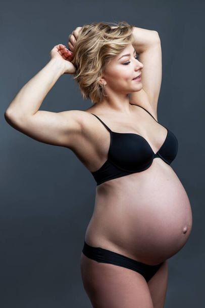 Pregnant woman in black underwear. Beautiful young smiling blonde woman keeps her hands behind her head. Happiness in anticipation of a baby. Gray background. Vertical. - Foto, Imagem