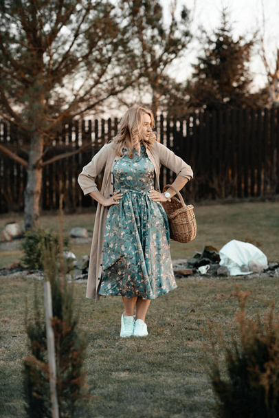 A cute girl in a long blue dress with a basket in her hands is walking in the park among the trees, spring has come. Walk in the park. Landscaping. Defocus. - Fotografie, Obrázek