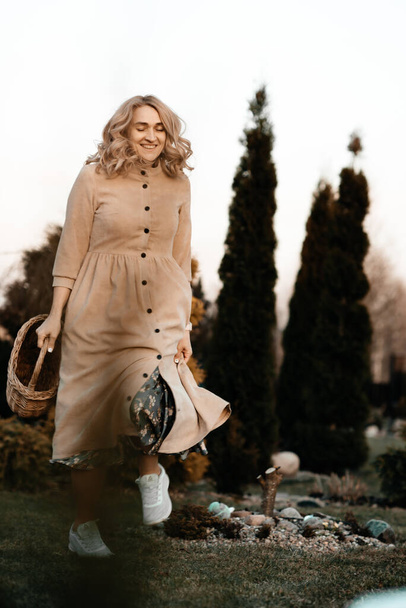 A sweet girl with blond curly hair in a long dress with a basket in her hands is walking in the park among the trees, spring has come. Walk in the park. Landscaping. Defocus. - Foto, afbeelding