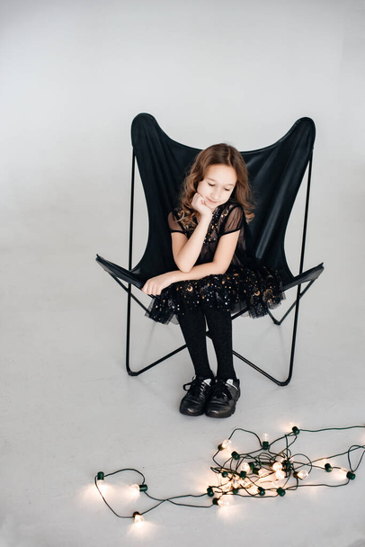 Happy little girl with dark curly hair in a black dress sits on an armchair in the studio enjoying life, the lights are blinking. The concept of children's style and fashion. Defocus. - Foto, afbeelding