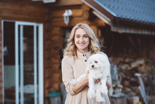 A blonde girl with curly hair in a long dress holds a small white dog in her hands while standing in the open air against the background of a wooden house. Pedigree dog. Pets concept. White puppy. - Fotografie, Obrázek