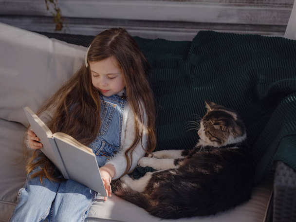 Autumn sunny weekend with cat at home. little girl reading a book on the veranda with pet, home education for children. stay at home, Children and Pets Relationships Concept - 写真・画像