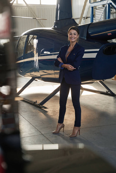 Joyous businesswoman posing for camera near hangared helicopter - 写真・画像