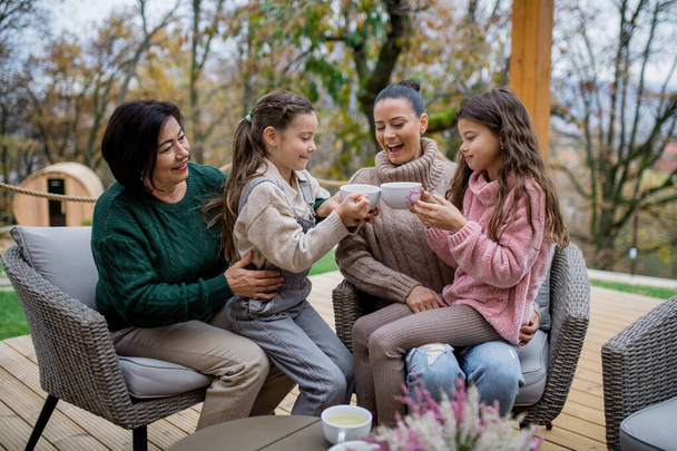 Two happy sisters with mother and grandmother sitting and drinking tea outdoors in patio in autumn. - Foto, imagen