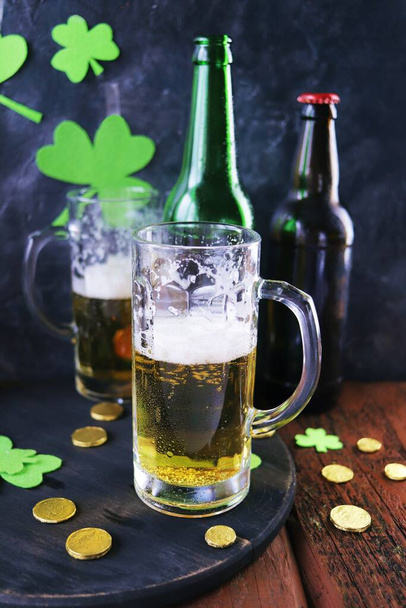 Patrick day, foamy beer in glass mugs and a bottle, gold coins on a wooden table, green shamrock on a dark background, party, congratulation, postcard - Foto, Bild