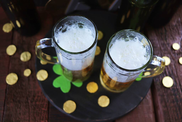 Patrick day, foamy beer in glass mugs and a bottle, gold coins on a wooden table, green shamrock on a dark background, party, congratulation, postcard - Photo, image