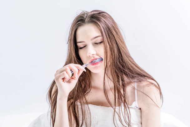 Sleepy brunette brushing her teeth while sitting on the bed in the white bedroom. The girl is engaged in oral hygiene. Wake up early in the morning - Photo, Image