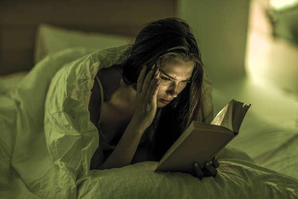 Beautiful woman reads a book intently under dim light in her bedroom before bed. Self-development. Relaxation. Literature. - Fotografie, Obrázek