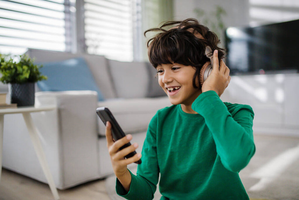 Happy multiracial boy with headphones and smartphone listening to music at home. - Photo, Image