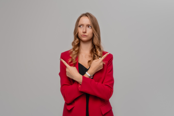 Indecisive blonde young woman pointing sideways, making tough decision, standing in red blazer against gray background - Zdjęcie, obraz